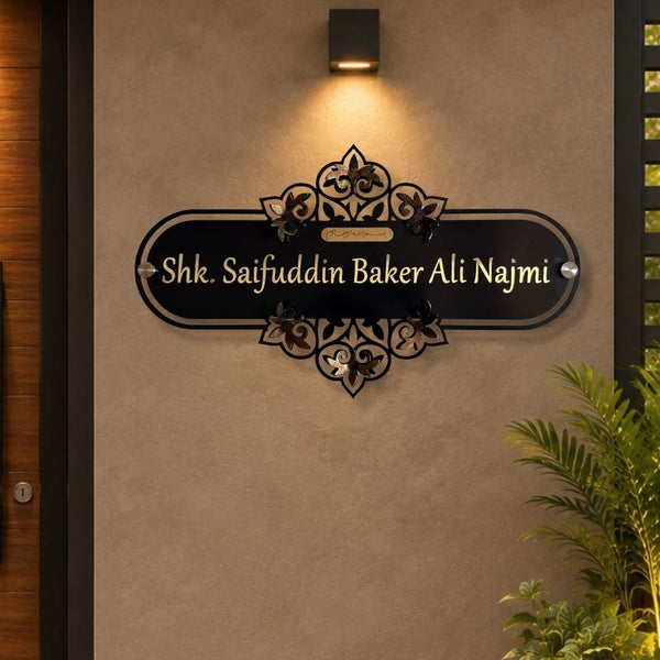 A custom black metal sign with ornate scrollwork and gold lettering reading 'Shk. Saifuddin Baker Ali Najmi', mounted on a textured beige wall under a black wall sconce.