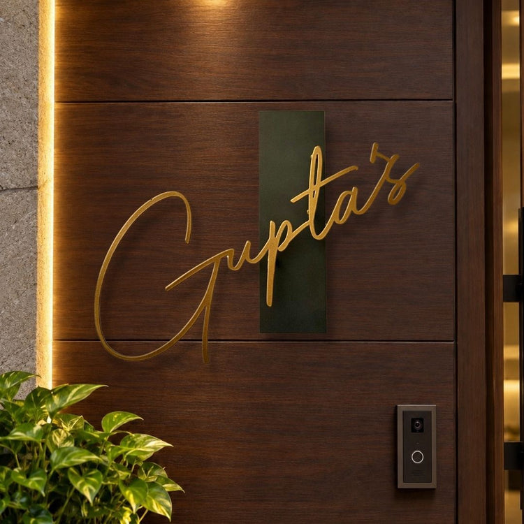A close-up of a dark wood door with a gold script sign reading 'Gupta's' mounted on a dark green rectangular plaque. Warm vertical lighting runs along the left edge, and a small plant and doorbell are visible in the foreground.

