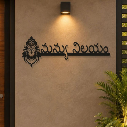 Black metal wall art featuring a stylized Hindu deity face and Telugu script mounted on a textured beige wall under a black outdoor sconce, with a green plant in the lower right corner.

