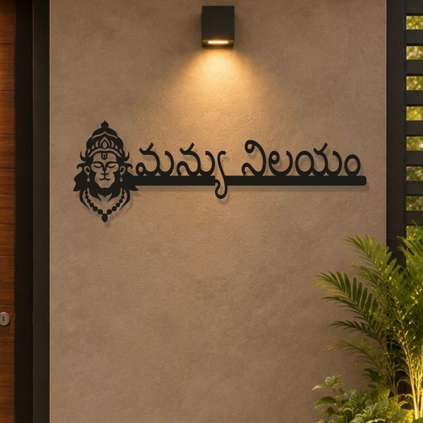 Black metal wall art featuring a stylized Hindu deity face and Telugu script mounted on a textured beige wall under a black outdoor sconce, with a green plant in the lower right corner.

