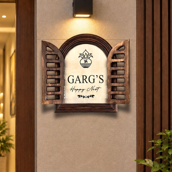 A decorative wall sign shaped like an open wooden shutter with a cream center panel reading “GARG’S Happy Meal” and a small flower emblem above. The sign is mounted on a beige wall under a lit black sconce, with warm wood trim and indoor plants visible around it.

