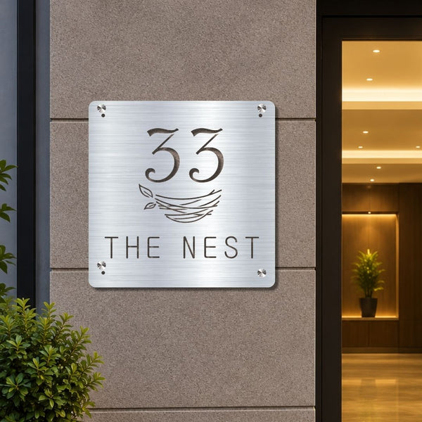 A square brushed-metal house number sign reading '33 THE NEST' mounted on a beige stone exterior wall, with a warmly lit building entrance visible to the right.

