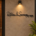 Black metal custom name wall sign reading 'Ritika & Sarang C-101' mounted on a textured beige wall under a warm wall sconce, with a plant visible in the lower right corner.

