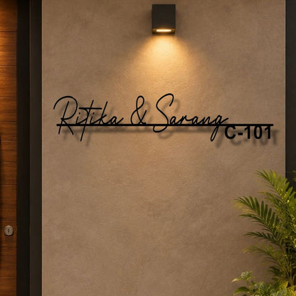 Black metal custom name wall sign reading 'Ritika & Sarang C-101' mounted on a textured beige wall under a warm wall sconce, with a plant visible in the lower right corner.

