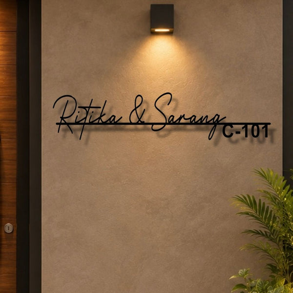 Black metal custom name wall sign reading 'Ritika & Sarang C-101' mounted on a textured beige wall under a warm wall sconce, with a plant visible in the lower right corner.

