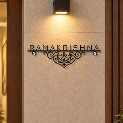 Black metal wall sign with the name 'RAMAKRISHNA' and an ornate scrollwork design, mounted on a beige textured wall under a black wall sconce.

