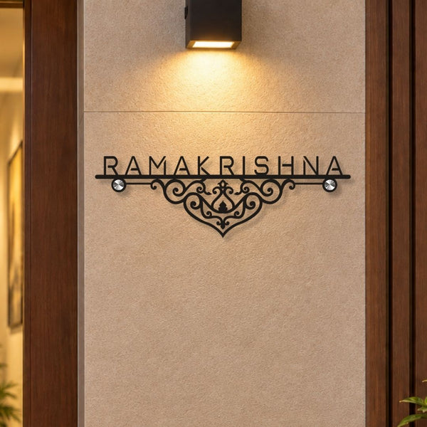 Black metal wall sign with the name 'RAMAKRISHNA' and an ornate scrollwork design, mounted on a beige textured wall under a black wall sconce.

