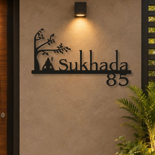 Black metal custom house number sign reading 'Sukhada 85' with a tree and tent silhouette, mounted on a beige stucco wall under a black outdoor sconce.

