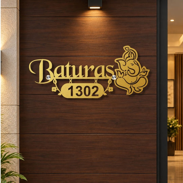 Gold-colored custom house number sign featuring the name 'Baturas' in cursive, the number '1302' in a badge, and a Ganesha illustration, mounted on a dark wood-paneled exterior door.


