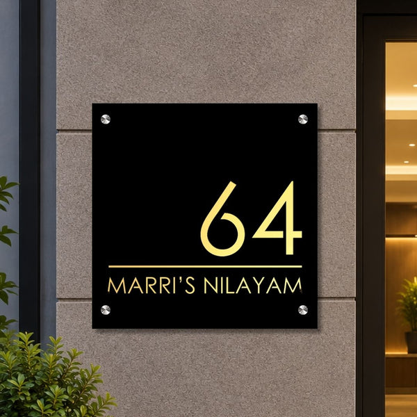 A black square custom house number plaque with gold lettering reading '64 MARRI'S NILAYAM', mounted on a textured gray stone wall.