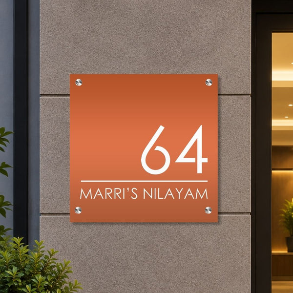An orange square acrylic house number sign mounted on a gray stone wall, featuring the white text '64' and 'MARRI'S NILAYAM' with small silver corner brackets.


