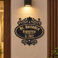 An oval black metal house number plaque with gold lettering reading 'Dr. Ravindra Uspariya A-302' and ornate scrollwork, mounted on a beige wall under a wall light.

