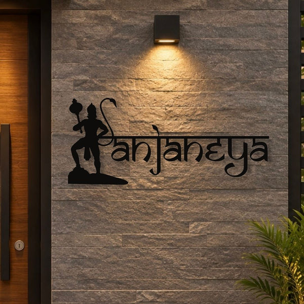Black metal wall art featuring the Devanagari name 'Sanjaneya' alongside a silhouette of Lord Hanuman, mounted on a gray stone-textured wall under a warm wall light.


