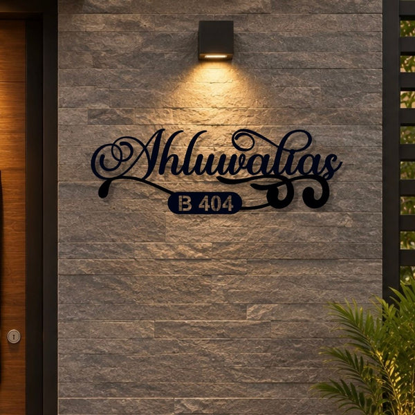 A black metal custom address sign reading 'Ahluwalias B 404' mounted on a gray stone wall under a warm outdoor light, with a wooden door on the left and green plants at the bottom right.

