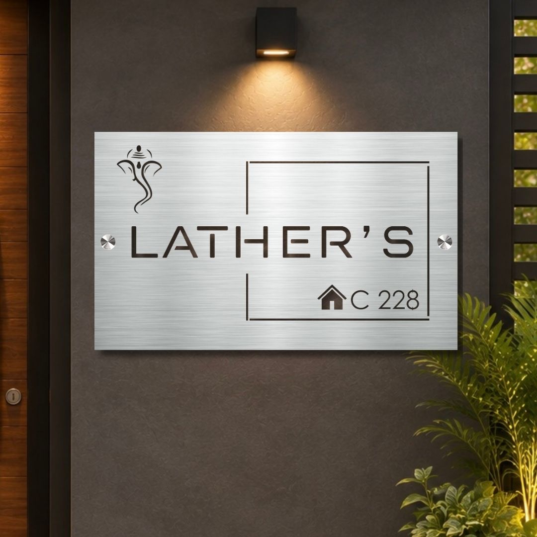 A rectangular brushed-metal house sign mounted on a dark gray wall, featuring the engraved text 'LATHER'S' and 'C 228' with a small house icon. A warm wall-mounted light above casts a glow on the sign, with plants and a wooden door visible at the edges.

