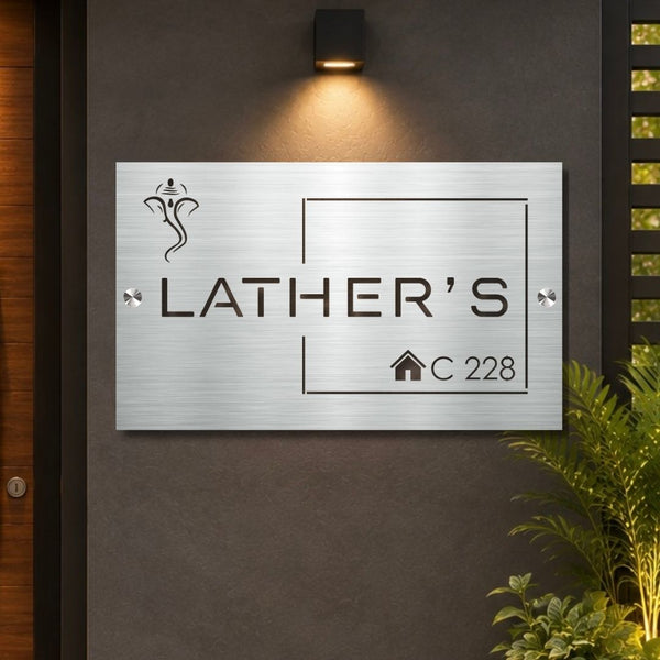 A rectangular brushed-metal house sign mounted on a dark gray wall, featuring the engraved text 'LATHER'S' and 'C 228' with a small house icon. A warm wall-mounted light above casts a glow on the sign, with plants and a wooden door visible at the edges.

