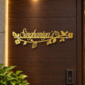 A personalized gold metal door sign reading 'Singhaniya's' mounted on a dark brown wooden door, decorated with leaves, flowers, and a small bird.

