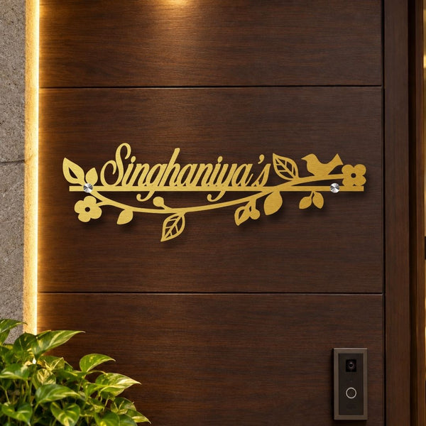A personalized gold metal door sign reading 'Singhaniya's' mounted on a dark brown wooden door, decorated with leaves, flowers, and a small bird.

