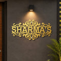 A decorative gold metal sign spelling 'SHARMA'S' with ornate scrollwork, mounted on a dark gray textured wall under a black wall sconce. A green plant is visible in the lower right corner.

