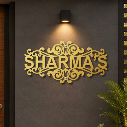 A decorative gold metal sign spelling 'SHARMA'S' with ornate scrollwork, mounted on a dark gray textured wall under a black wall sconce. A green plant is visible in the lower right corner.

