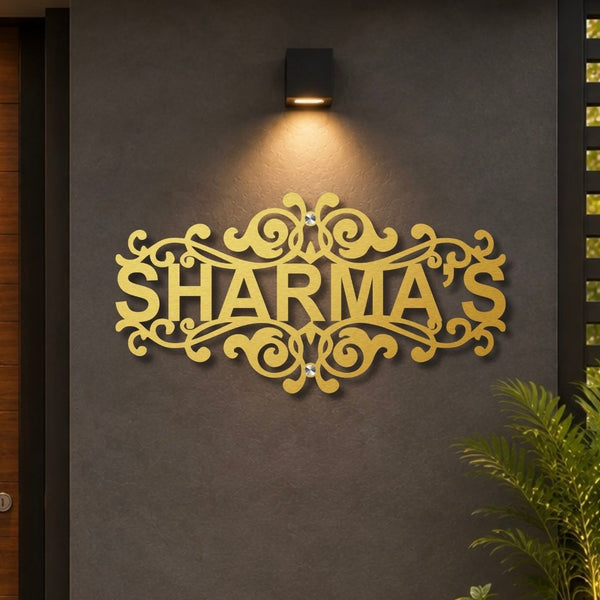 A decorative gold metal sign spelling 'SHARMA'S' with ornate scrollwork, mounted on a dark gray textured wall under a black wall sconce. A green plant is visible in the lower right corner.

