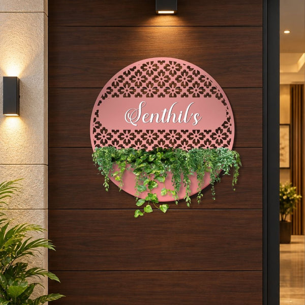 A pink circular decorative wall sign with a laser-cut floral pattern and the script text 'Senthil's' in the center, mounted on a dark wood-paneled wall. Green trailing plants are placed in the lower cutout of the sign, and warm wall lights and a doorway are visible in the background.

