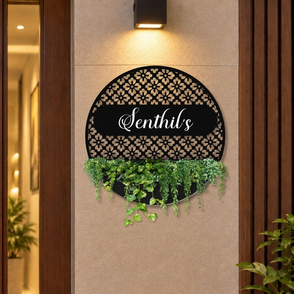 A decorative circular black wall sign with a floral cutout pattern, the name 'Senthil's' in white script across the center, and trailing green ivy hanging from the bottom, mounted on a beige wall between wooden door frames.

