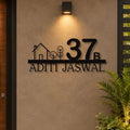 Custom black metal wall art featuring the number 37, a house outline, trees, and the name 'ADITI JASWAL' mounted on a beige stucco wall under a wall sconce.

