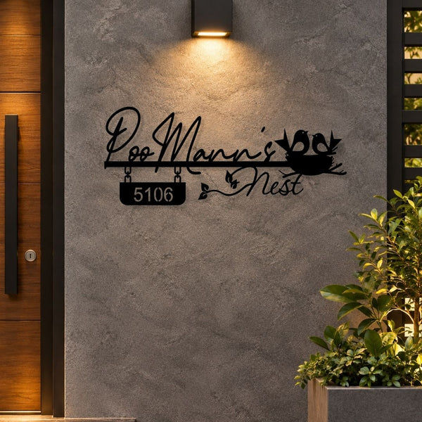 Black metal personalized wall art featuring the text 'Doo Mann's Nest' with a hanging house number plaque '5106' and a bird nest motif, mounted on a textured gray wall in an outdoor entryway setting.

