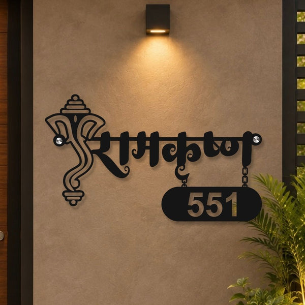 Black metal wall art featuring the word 'Raksha' in Devanagari script with a hanging plaque displaying '551' and a decorative Ganesha motif, mounted on a beige stucco wall under a wall light.

