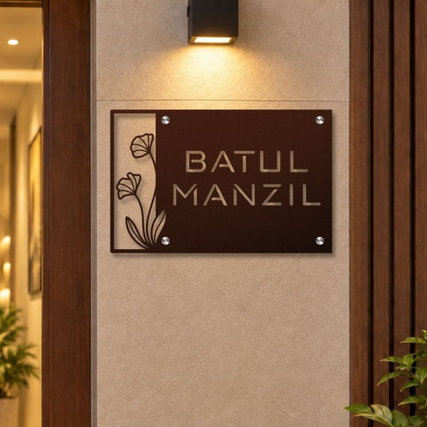 A rectangular dark brown metal sign with the words 'BATUL MANZIL' in gold lettering, mounted on a beige textured wall under a black wall sconce. The left side of the sign features a decorative cutout floral design.

