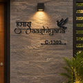 Black metal house number and name sign featuring Devanagari script, a dove silhouette, and house number C-1303, mounted on a gray stone wall under a warm light.

