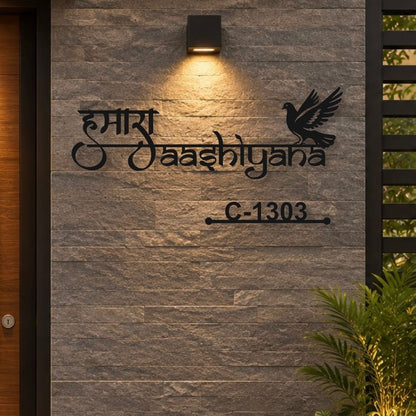 Black metal house number and name sign featuring Devanagari script, a dove silhouette, and house number C-1303, mounted on a gray stone wall under a warm light.

