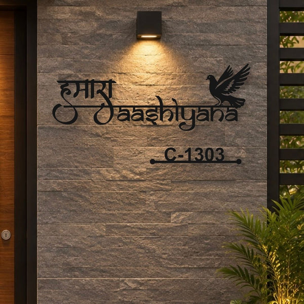 Black metal house number and name sign featuring Devanagari script, a dove silhouette, and house number C-1303, mounted on a gray stone wall under a warm light.

