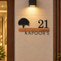 Custom metal house number sign reading '21 KAPOOR'S' with a black tree silhouette on a wooden plank, mounted on a beige wall next to a dark wood door frame under a wall light.

