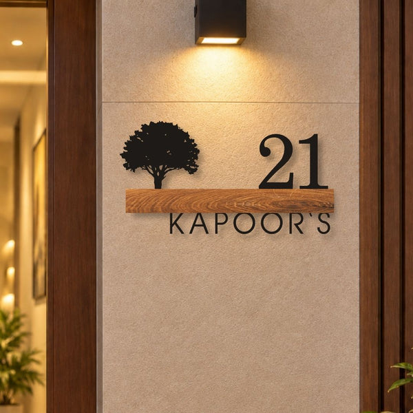 Custom metal house number sign reading '21 KAPOOR'S' with a black tree silhouette on a wooden plank, mounted on a beige wall next to a dark wood door frame under a wall light.


