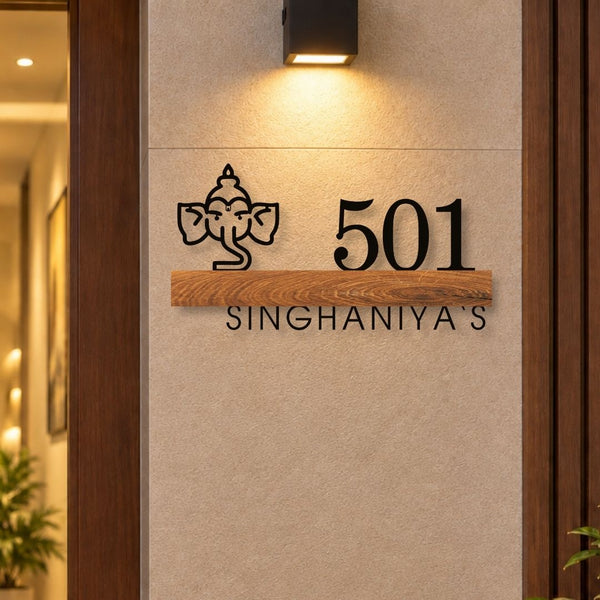 A custom black metal house number sign featuring a Ganesha icon, the number 501, and the name 'SINGHANIYA'S' mounted on a beige textured wall with a warm wall sconce above.

