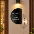 Modern metal house number and name sign mounted on a beige wall, featuring a black circular nameplate with gold lettering and a separate vertical silver number plate next to it.

