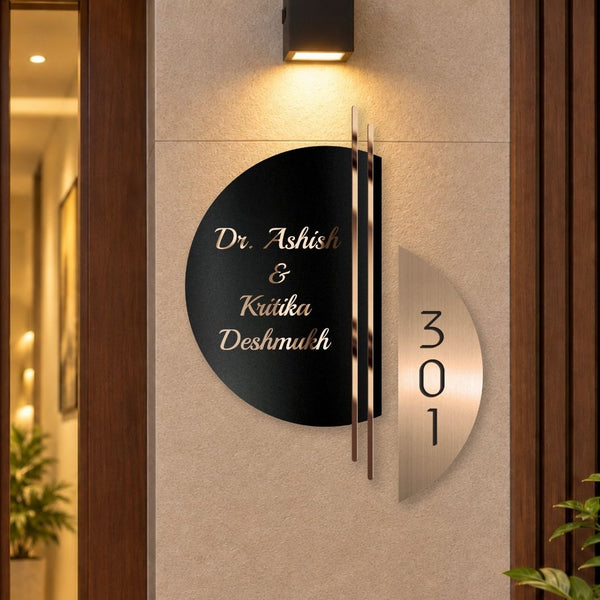 Modern metal house number and name sign mounted on a beige wall, featuring a black circular nameplate with gold lettering and a separate vertical silver number plate next to it.

