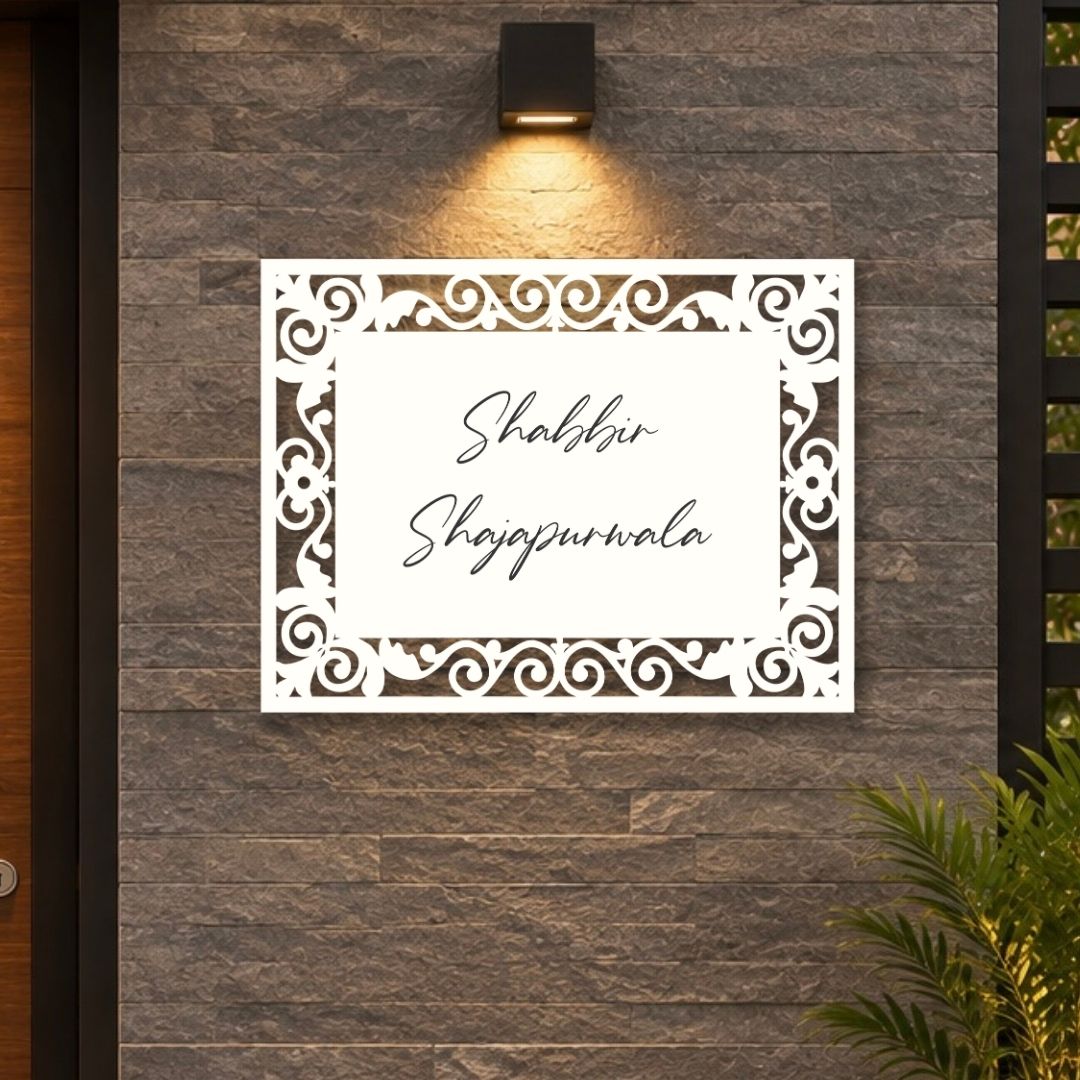 A white rectangular laser-cut sign with ornate scrollwork borders and the Urdu text 'Shabbir Shajpwrala' in black script, mounted on a dark gray stone wall under a black wall sconce.

