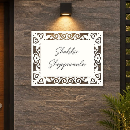 A white rectangular laser-cut sign with ornate scrollwork borders and the Urdu text 'Shabbir Shajpwrala' in black script, mounted on a dark gray stone wall under a black wall sconce.

