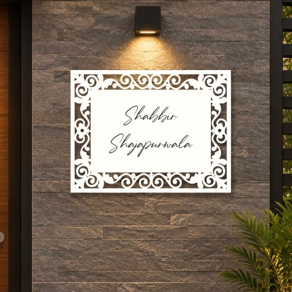 A white rectangular laser-cut sign with ornate scrollwork borders and the Urdu text 'Shabbir Shajpwrala' in black script, mounted on a dark gray stone wall under a black wall sconce.

