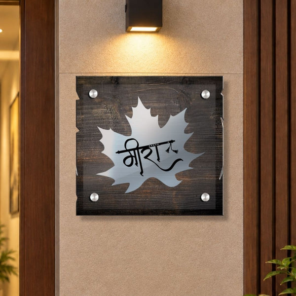 A square dark wood plaque with a silver maple leaf shape and the word 'MIRAZ' in black script, mounted on a beige wall under a black wall light.

