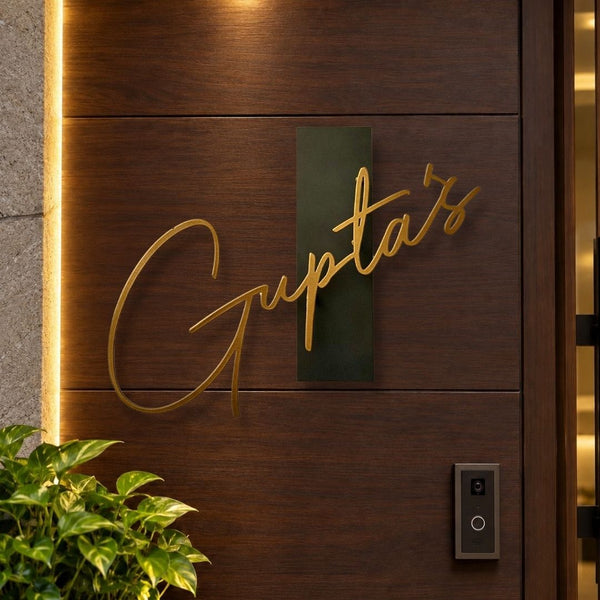A close-up of a dark wood door with a gold script sign reading 'Gupta's' mounted on a dark green rectangular plaque. Warm vertical lighting runs along the left edge, and a small plant and doorbell are visible in the foreground.

