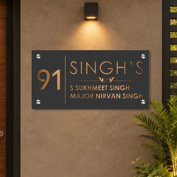 A custom rectangular black metal house number sign with gold lettering, mounted on a textured beige wall under a wall light. The sign displays the number 91 and the name 'SINGH'S' with additional custom text below, flanked by decorative silver rivets.

