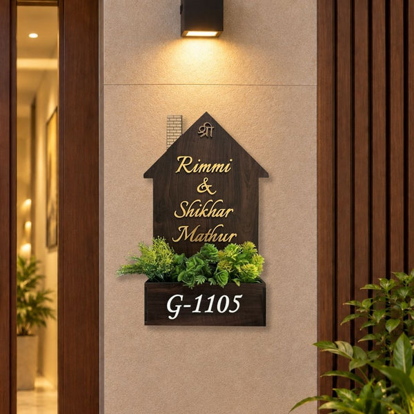 A personalized house number sign mounted on a beige wall. The sign is shaped like a dark brown wooden house with gold-colored text reading 'Rimmi & Shukhar Mathur' and a small monogram 'SM' at the top.


