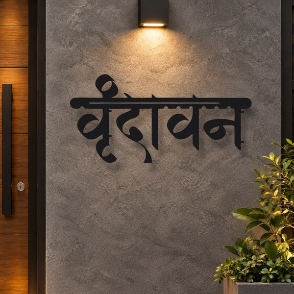 Black metal wall art featuring the Devanagari word 'Veedavani' mounted on a textured gray concrete wall, illuminated by a warm overhead light. A dark wooden door is visible on the left, and green potted plants are in the lower right corner.
