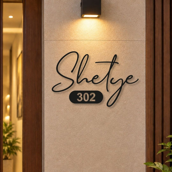 Black metal script house number sign reading 'Shetye 302' mounted on a beige stone wall, with a warm wall sconce above and a doorway with wood trim on the left.