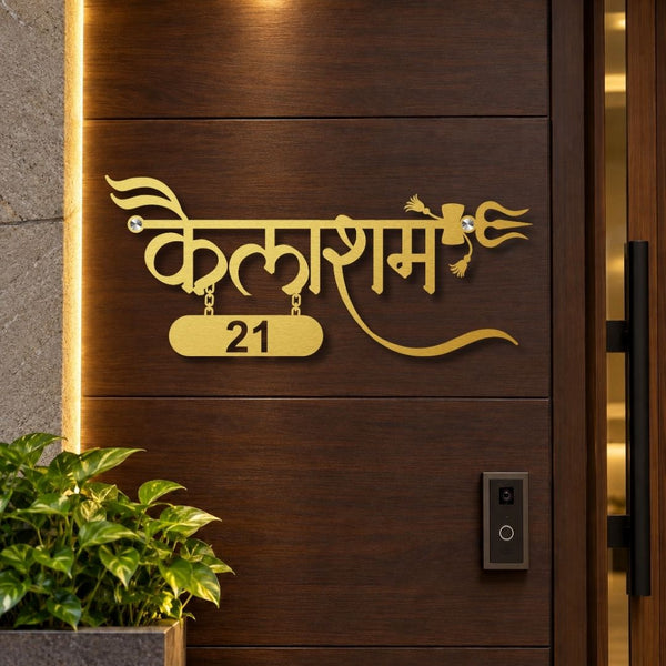 Gold-colored metal Hindi nameplate with the number 21 on a dark brown wooden door, shown in a real-world entryway setting with a plant and doorbell visible