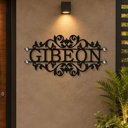 Black metal personalized name sign reading 'GIBEON' with ornate scrollwork and small clear stone accents, mounted on a beige stucco wall under a wall light.